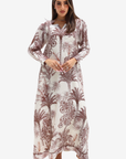 A woman modeling a long-sleeved maxi dress with a brown palm tree and floral toile pattern on a cream background, set against a plain light gray studio background.