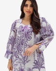 A woman modeling a loose, long-sleeve tunic dress with a purple botanical and palm tree print on a white background, featuring a purple tie-neck detail.
