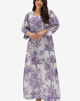 A woman modeling a long-sleeved maxi dress with a purple palm tree toile pattern on a white background, set against a plain light gray studio backdrop.
