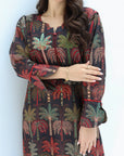 A woman wearing a long-sleeve tunic dress with a tropical palm tree print on a dark background. The dress features red, green, and beige palm trees with red piping details along the collar, cuffs, and front button placket.