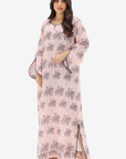 A woman modeling a long light pink kaftan dress with a gray palm tree pattern, shown against a plain light gray background.