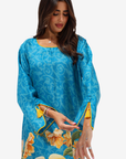 A woman wearing a bright blue tunic dress with a subtle tonal swirl pattern, yellow floral details, and two yellow birds near the hem, shown against a plain light gray background.