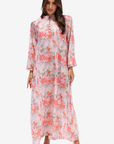 A woman modeling a long-sleeved, high-neck maxi dress with a pink, coral, and green floral print on a light background, set against a plain light gray studio background.