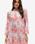 A woman modeling a long-sleeved, high-neck tunic dress with a pink and coral floral print on a light background, set against a plain light gray studio backdrop.