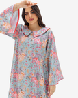A woman wearing a long-sleeved kaftan dress with a light blue-gray base and a colorful floral print in pink, orange, and purple, featuring a large Peter Pan collar and red piping details.
