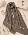 A taupe gray wool scarf with silver rhinestone borders, displayed in a flat-lay arrangement with its branded cream packaging box, drawstring pouch, and hanging tag on a light beige background.