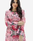 A woman modeling a pink long-sleeve tunic dress with a red and green floral pattern and black lace trim details, set against a plain light gray background.