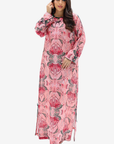 A woman modeling a long-sleeved pink maxi dress with a red and green floral pattern, lace-up side details, and a round neckline, set against a plain light gray background.