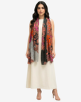A woman modeling a long, lightweight scarf with a vibrant multicolor paisley pattern in orange, pink, black, and gray, worn over a cream-colored dress against a light gray studio background.