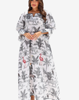 A woman modeling a long white maxi dress with a black and gray animal and palm tree print, featuring pink flamingo accents, set against a plain light gray background.