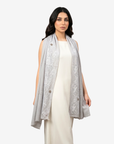 A woman modeling a light gray sleeveless shawl with white floral embroidery and decorative buttons, worn over a cream-colored dress against a plain light gray background.