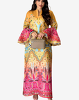 A woman modeling a long yellow and pink printed dress with bell sleeves and a geometric pattern, holding a small beige handbag against a light gray background.