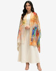 A woman modeling a cream-colored dress with a vibrant, multicolored abstract painterly shawl draped over her shoulders, set against a plain light gray background.
