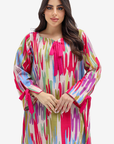 A woman modeling a long-sleeved, loose-fitting tunic dress with a vibrant multicolor abstract print. The dress features a bright pink tie-neck bow and matching pink tie details on the cuffs, set against a plain light gray studio background.