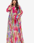 A woman modeling a long, flowing abaya dress with a vibrant multicolor ikat pattern in pink, blue, green, and yellow on a cream base, set against a plain light gray background.