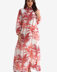 A woman modeling a long-sleeved, collared maxi dress with a red palm tree print on a white background, set against a plain light gray studio backdrop.