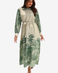 A woman modeling a long-sleeve midi dress with a cream base and green tropical leaf print, featuring a matching fabric tie belt at the waist, set against a plain light gray background.