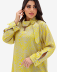 A woman modeling a yellow and gray patterned long-sleeve tunic dress with a high collar, bow detail, and button cuffs against a light gray studio background.
