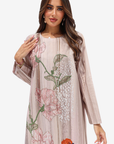 A woman modeling a long-sleeved, floor-length tunic dress in a light beige-pink color, decorated with large pink and orange floral embroidery and green leaf details on a solid white background.