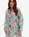 A woman modeling a long-sleeve V-neck tunic dress with a colorful seashell and starfish print on a cream background, featuring green tie details at the cuffs.