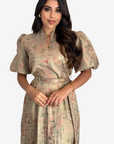 A woman modeling a short-sleeved midi dress with puff sleeves, a V-neckline, and a matching waist belt. The dress features a shimmering metallic gold base with a subtle abstract print in muted pink and green tones.