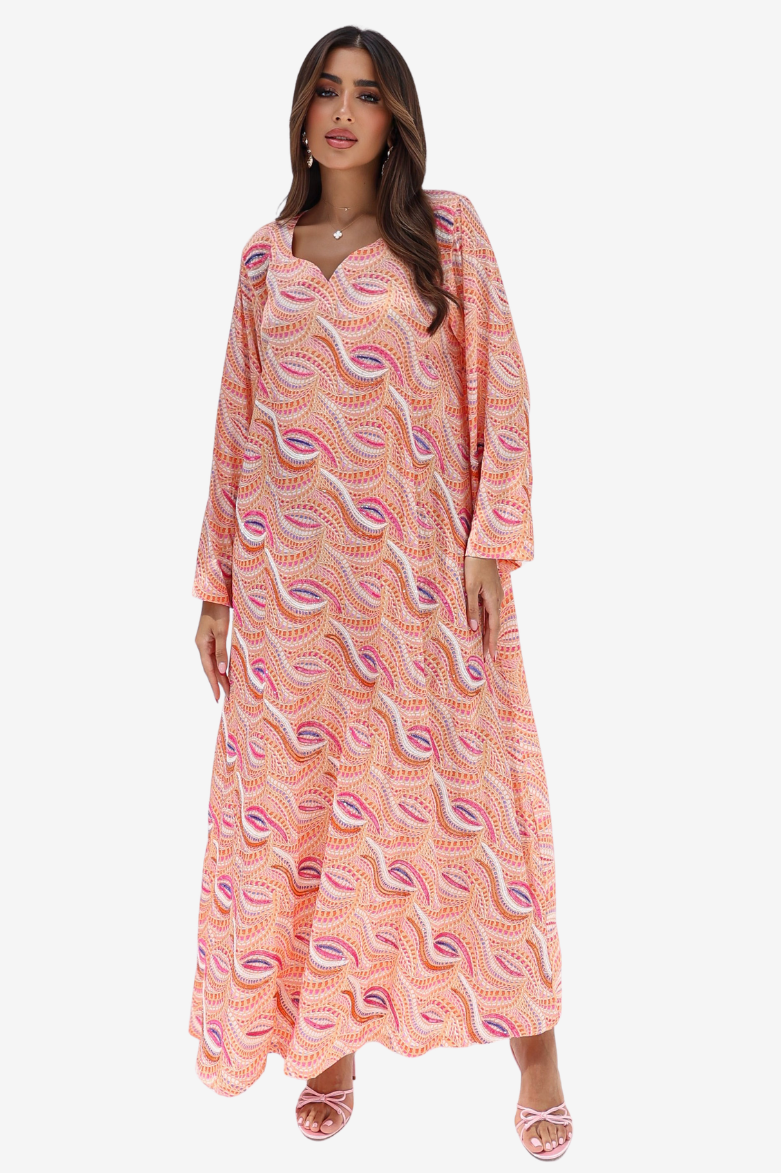 A woman modeling a long-sleeve, floor-length kaftan dress with a peach and pink paisley pattern, set against a plain light gray background.