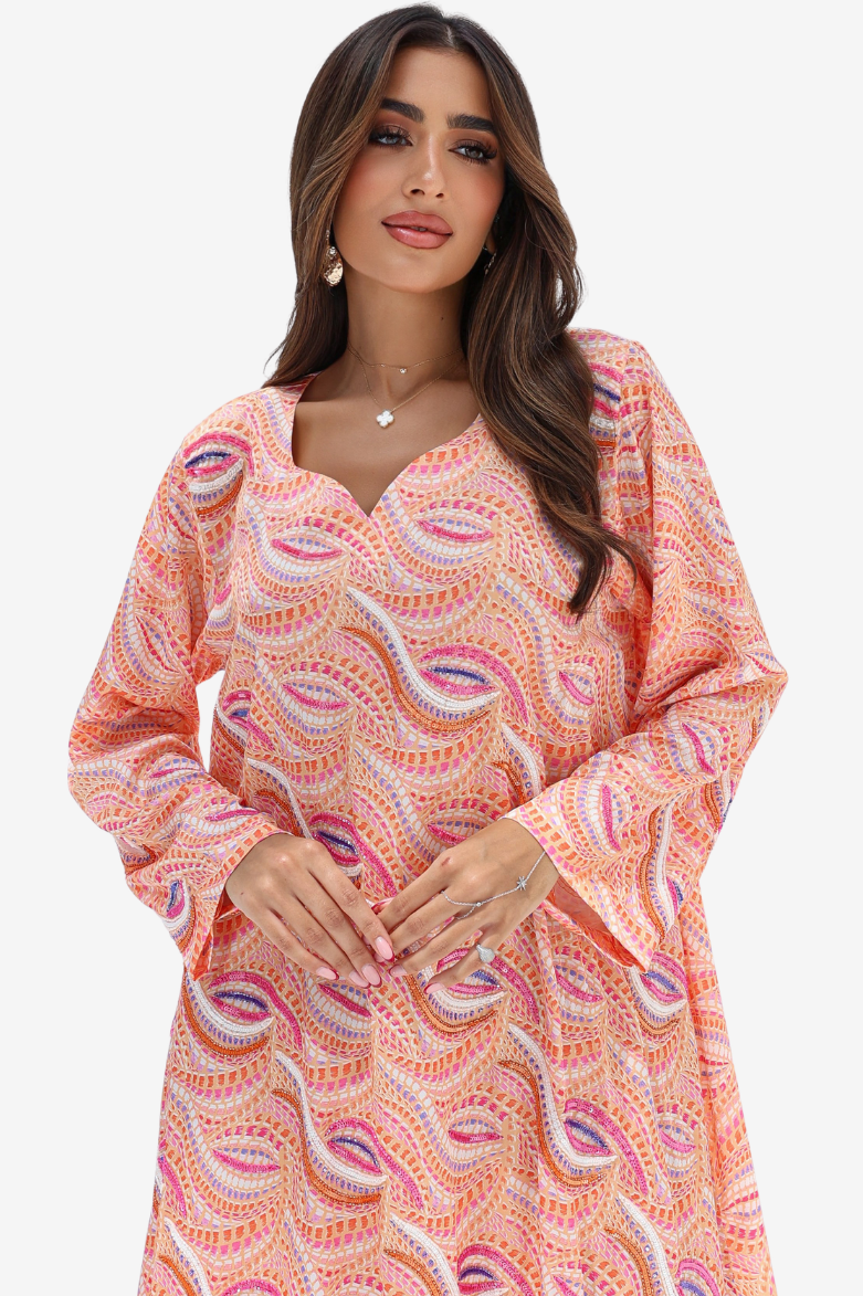 A woman wearing a peach-colored long-sleeve tunic dress with a pink, purple, and white paisley pattern, photographed against a solid light gray background.