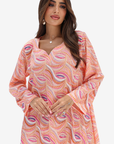 A woman wearing a peach-colored long-sleeve tunic dress with a pink, purple, and white paisley pattern, photographed against a solid light gray background.