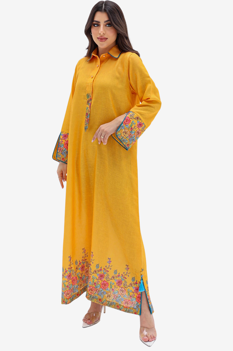 A woman modeling a long yellow kaftan dress with colorful floral embroidery on the cuffs and hem, set against a plain light gray background.