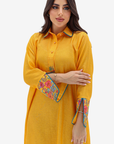 A woman modeling a mustard yellow long-sleeve kurta with colorful floral embroidery on the cuffs and collar, set against a plain light gray background.