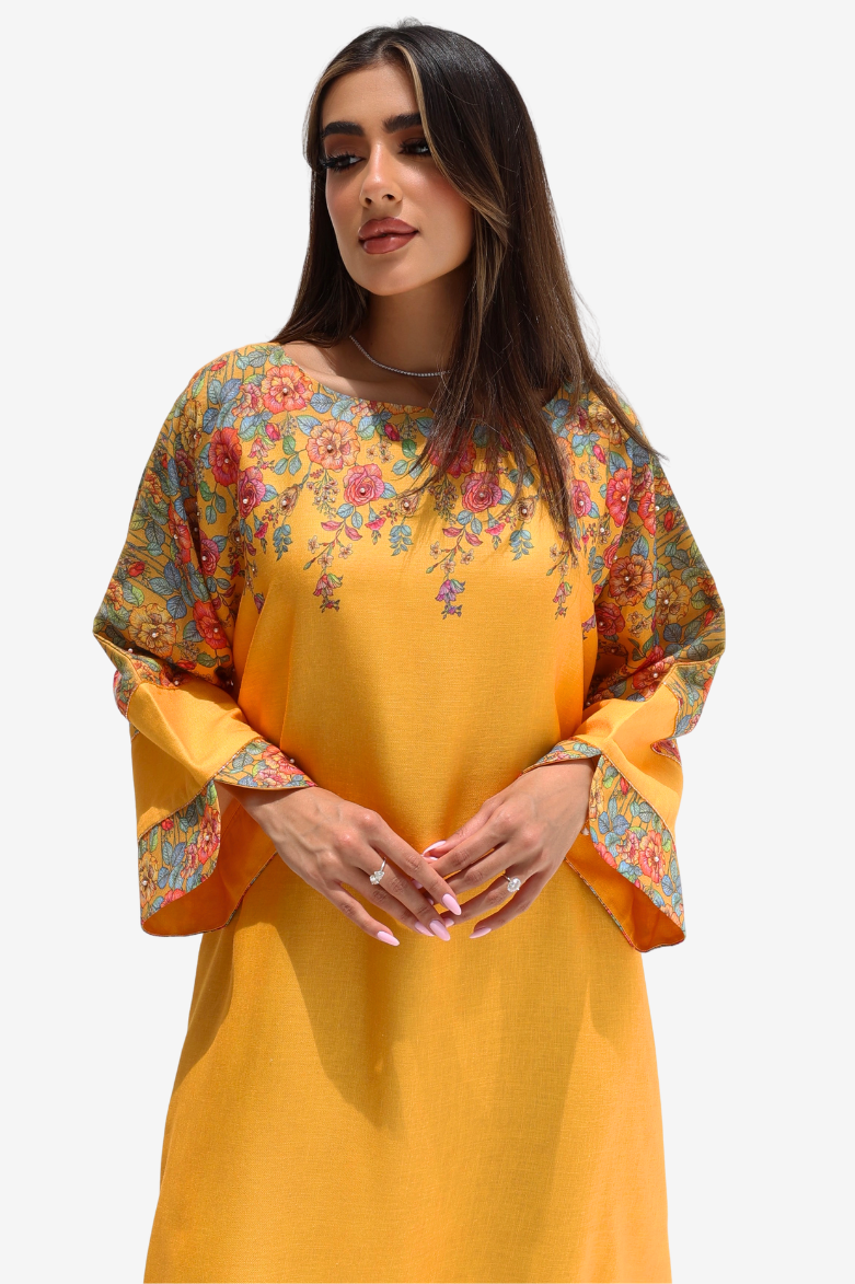 A woman wearing a mustard yellow tunic dress with colorful floral embroidery on the yoke and wide bell sleeves, set against a plain light gray background.