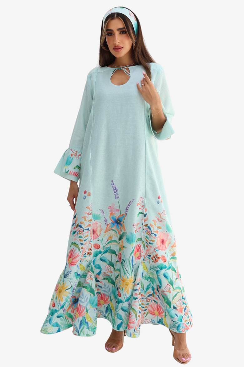 A woman modeling a long, flowing kaftan dress in mint green with a colorful floral and butterfly print concentrated along the hem, styled with a matching headband against a plain white background.