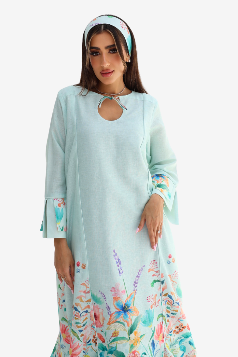 A woman modeling a light mint green maxi dress with a colorful floral print at the hem and cuffs, paired with a matching floral headband, against a plain white background.