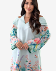A woman modeling a light blue off-the-shoulder tunic dress with vibrant multicolor floral patterns on the sleeves and hem, set against a plain light gray background.