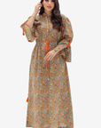 A woman modeling a long-sleeved, ankle-length floral maxi dress with a brown base and orange tie details, set against a plain light gray background.