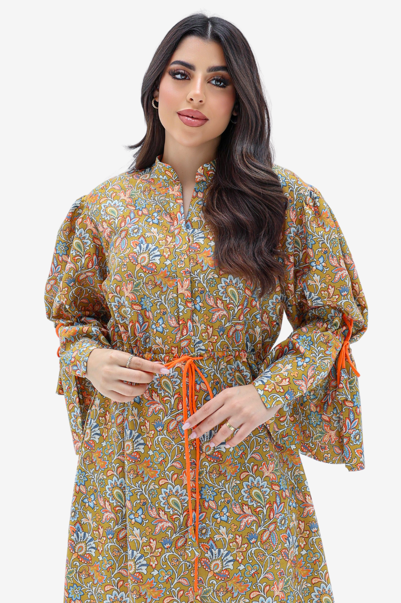 A woman modeling a long-sleeved, high-neck tunic dress with a dense multicolor paisley pattern on a mustard-yellow base, featuring an orange drawstring belt and orange tie details on the sleeves, set against a plain light gray background.