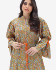 A woman modeling a long-sleeved, high-neck tunic dress with a dense multicolor paisley pattern on a mustard-yellow base, featuring an orange drawstring belt and orange tie details on the sleeves, set against a plain light gray background.