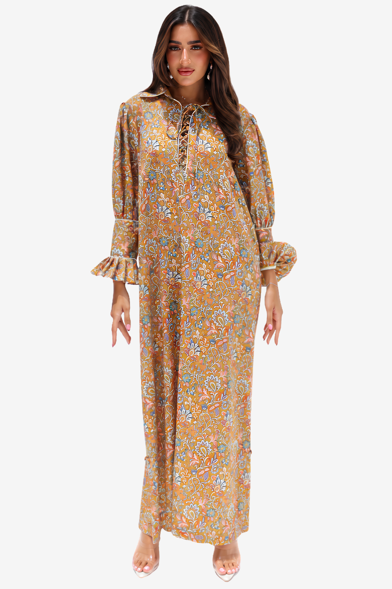 A woman modeling a long-sleeved maxi dress with a bohemian floral print in mustard yellow, orange, and blue. The dress features a lace-up neckline, ruffled cuffs, and a flowing silhouette, shown against a plain light gray background.