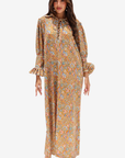 A woman modeling a long-sleeved maxi dress with a bohemian floral print in mustard yellow, orange, and blue. The dress features a lace-up neckline, ruffled cuffs, and a flowing silhouette, shown against a plain light gray background.