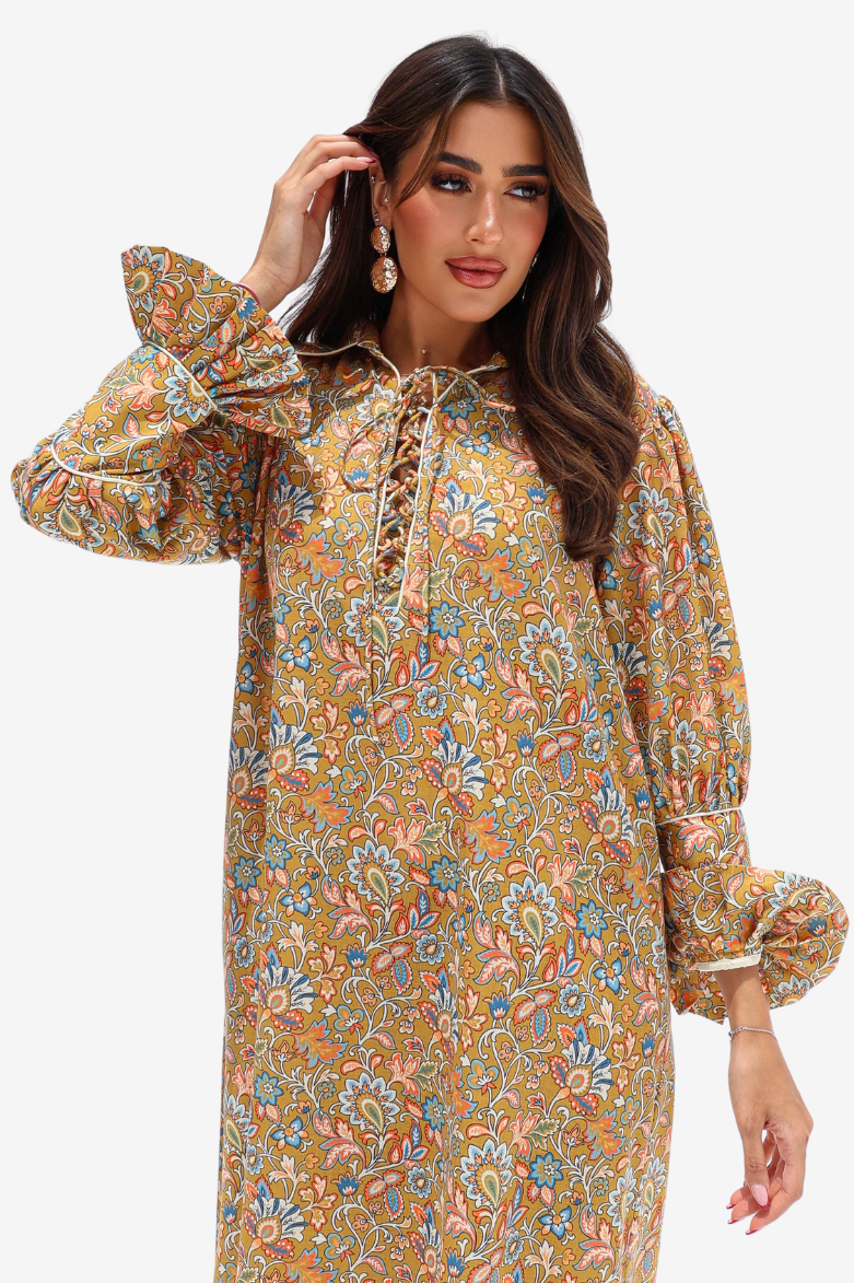 A woman modeling a long-sleeve bohemian-style tunic dress with a dense paisley floral print in mustard yellow, orange, blue, and green, featuring a lace-up neckline and ruffled cuffs.