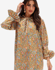 A woman modeling a long-sleeve bohemian-style tunic dress with a dense paisley floral print in mustard yellow, orange, blue, and green, featuring a lace-up neckline and ruffled cuffs.