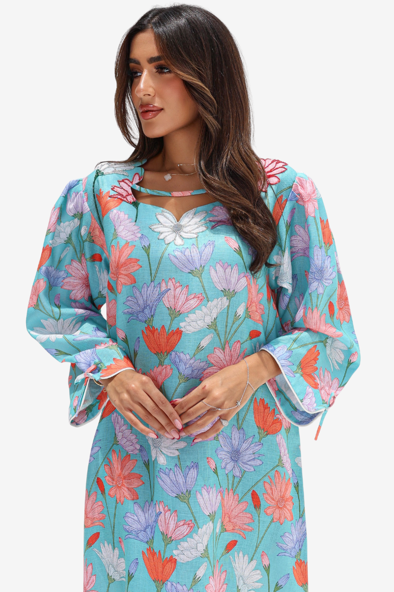 A woman wearing a long-sleeve turquoise floral dress with pink, purple, and white flowers, featuring a keyhole neckline with tie details and ruffled sleeves, set against a plain light gray background.