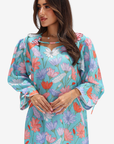 A woman wearing a long-sleeve turquoise floral dress with pink, purple, and white flowers, featuring a keyhole neckline with tie details and ruffled sleeves, set against a plain light gray background.