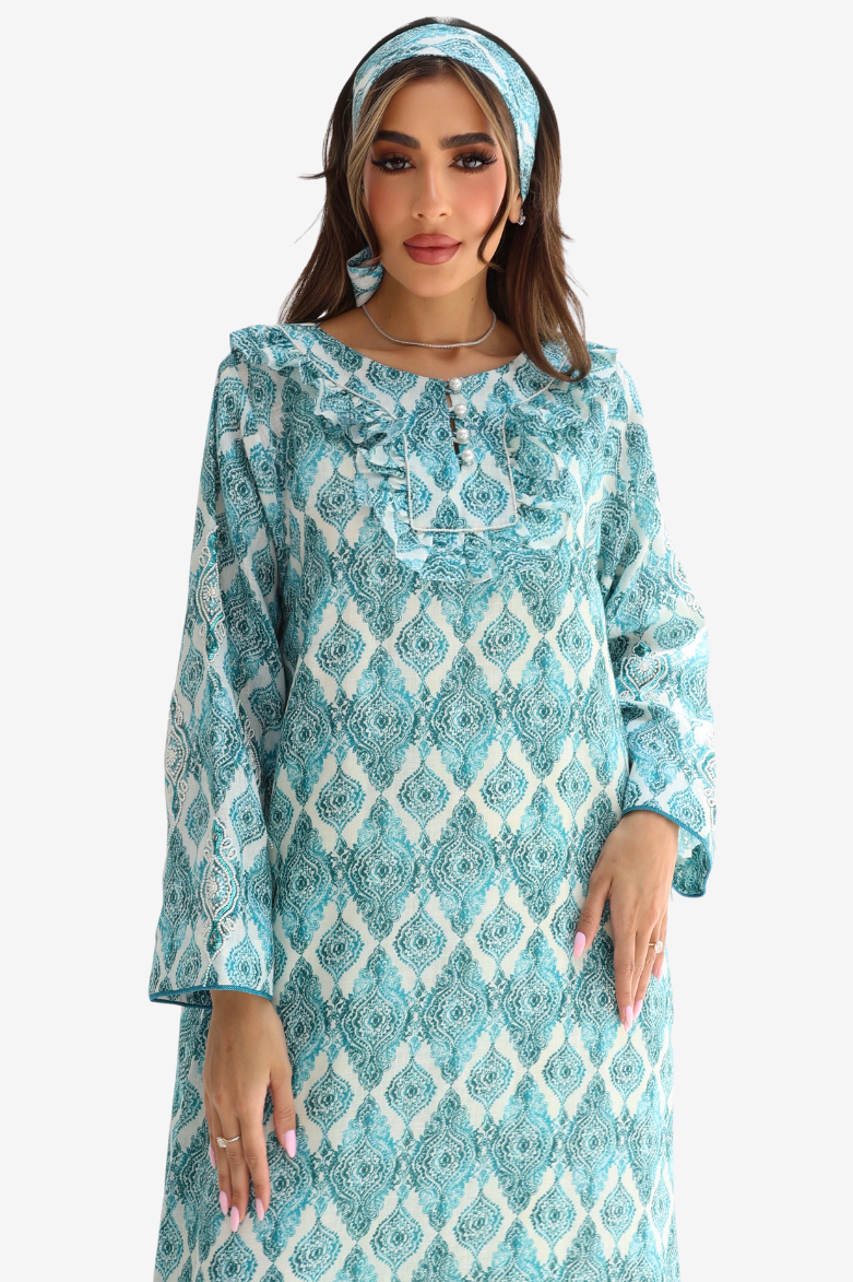 A woman wearing a long-sleeved turquoise and white patterned tunic dress with a ruffled collar, paired with a matching headband, set against a plain light gray background.