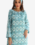 A woman wearing a long-sleeved turquoise and white patterned tunic dress with a ruffled collar, paired with a matching headband, set against a plain light gray background.
