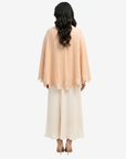 A woman shown from the back wearing a peach-colored lace-trimmed cape-style top over a long cream dress, against a plain light gray background.
