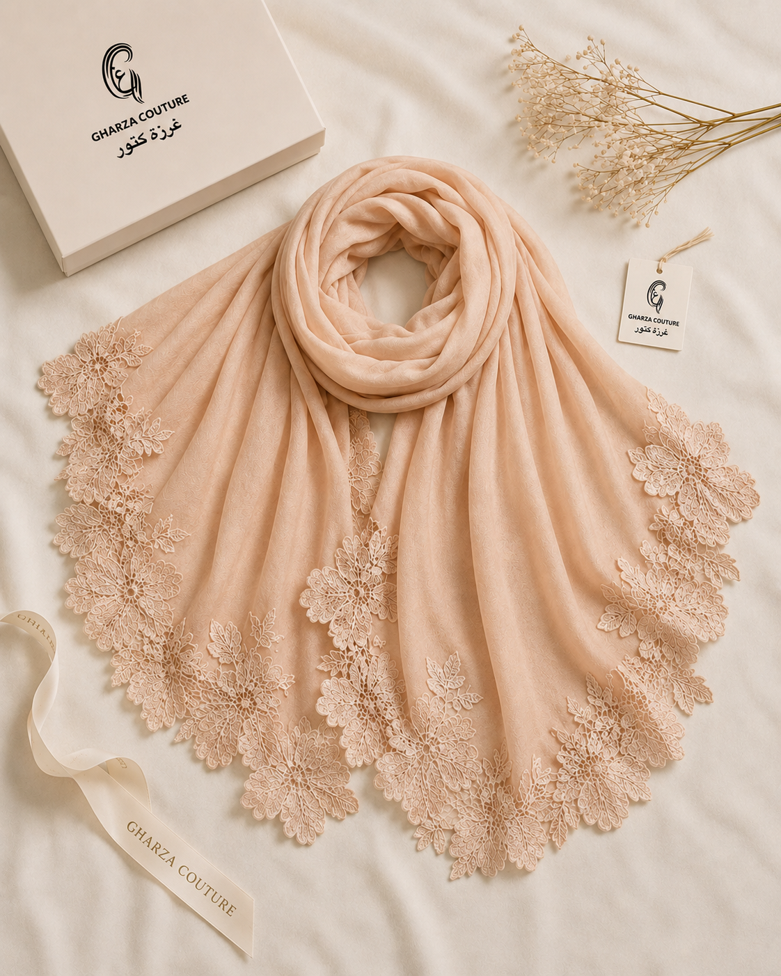 A peach-colored shawl scarf with intricate floral lace trim, elegantly draped on a white fabric background alongside a branded white box, a ribbon, and dried flowers. The brand 'GAMZA COUTURE' is visible on the packaging and ribbon.