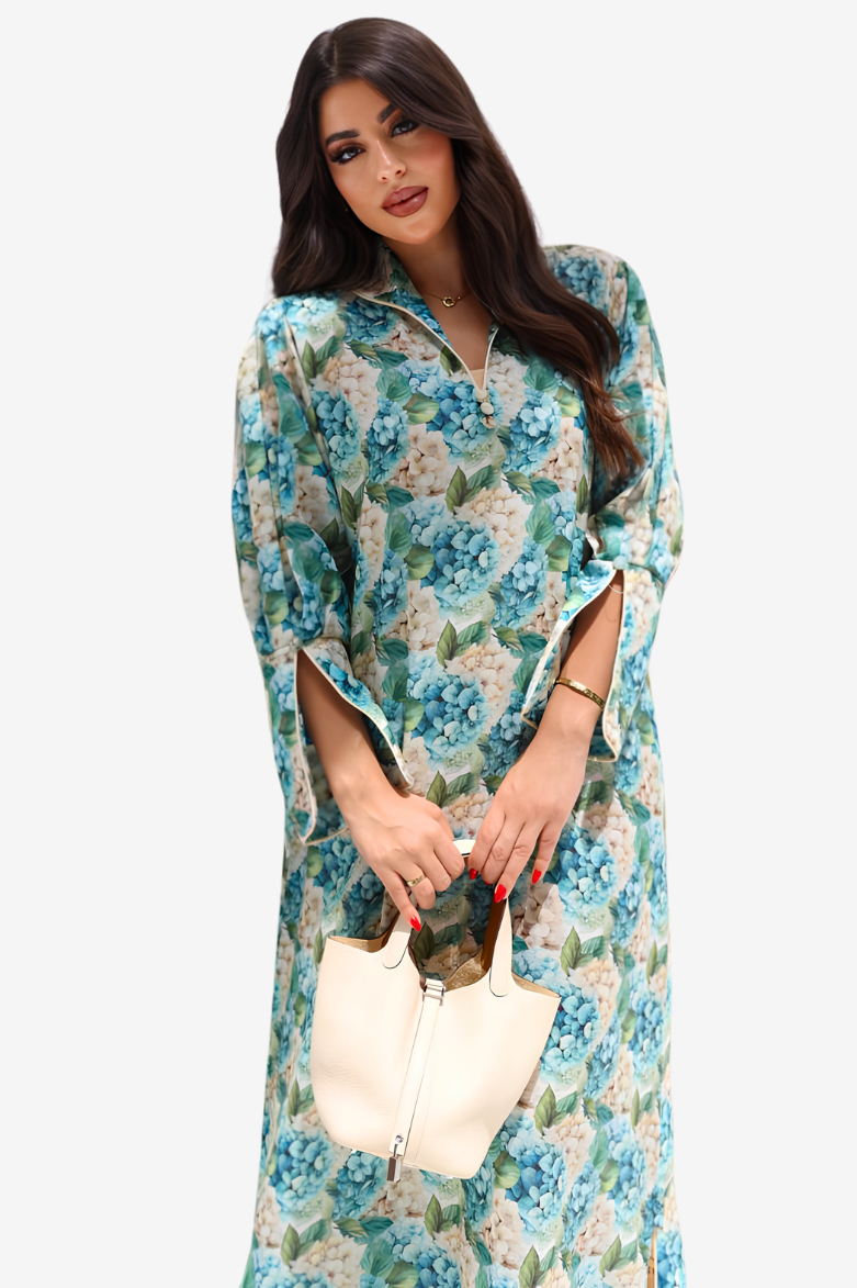 A woman modeling a long-sleeve floral maxi dress with a blue, green, and beige floral pattern, holding a cream-colored handbag against a light gray background.