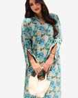 A woman modeling a long-sleeve floral maxi dress with a blue, green, and beige floral pattern, holding a cream-colored handbag against a light gray background.