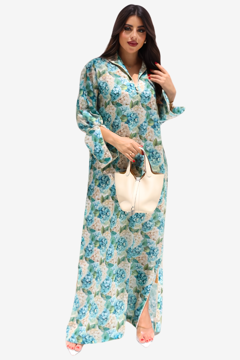 A woman modeling a long-sleeved, floor-length floral maxi dress with a high slit, holding a cream-colored handbag against a solid light gray background.